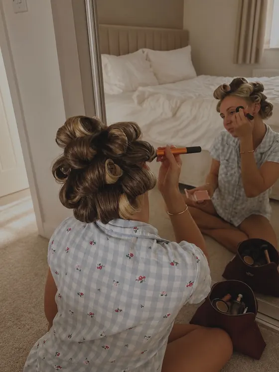 A set updo photographed in a bedroom while a friend does her makeup behind